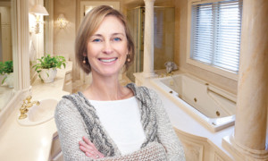 Smiling woman stands with arms crossed in a marble bathroom with a freestanding tub and columns behind her